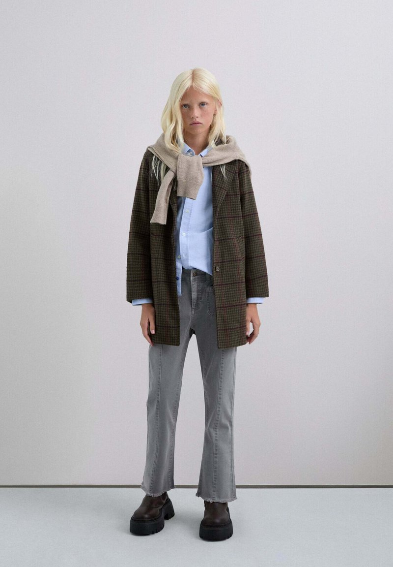 Jongeman met lange blonde haren, gekleed in een bruin geruite jas, een blauwe shirt, grijze versleten jeans, stevige zwarte laarzen en een beige trui op zijn schouders.