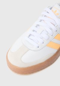 Biely kožený tenisk s jemne oranžovými pruhmi, semišovou špičkou, bielymi šnúrkami a textúrovanou gumovou podrážkou. Prítomný je minimálny branding.