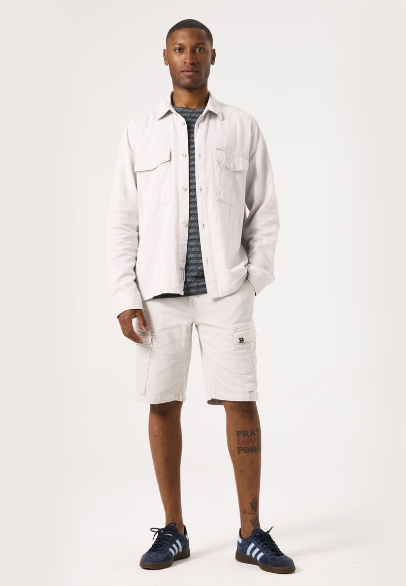 Chemise boutonnée gris clair par-dessus une chemise rayée bleu marine, associée à un short cargo beige et des baskets bleu foncé avec des accents blancs.