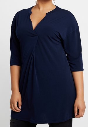 Woman wearing a navy blue tunic with three-quarter sleeves and a twisted front detail, paired with dark pants.