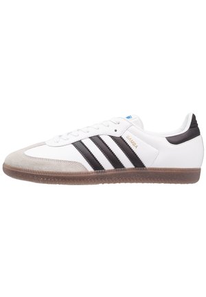 Witte Adidas Samba sneaker met zwarte strepen, suède neuskap, bruine zool en gouden "SAMBA" tekst aan de zijkant.