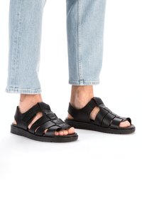 Sandales en cuir noir avec un design tressé, des côtés ouverts et une semelle en caoutchouc plate. Le matériau supérieur a une texture lisse et des accents minimaux.