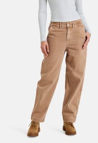 Pantalones ligeros de color marrón claro, holgados y con un tejido texturizado, que cuentan con un cierre de botón y una cinturilla elástica. Combinados con calzado resistente de color beige.