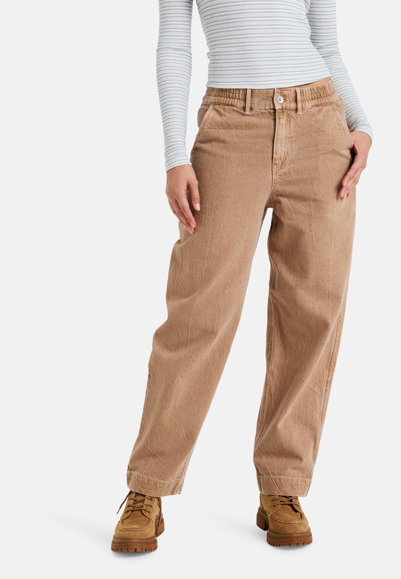 Pantalones ligeros de color marrón claro, holgados y con un tejido texturizado, que cuentan con un cierre de botón y una cinturilla elástica. Combinados con calzado resistente de color beige.
