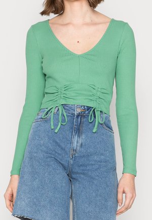 Green ribbed long-sleeve top with V-neck, gathered detail at the waist, and adjustable ties. Paired with high-waisted denim shorts.