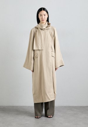 Beige long coat with a loose silhouette, featuring an attached scarf, two front pockets, and wide sleeves, paired with dark, wide-leg trousers.