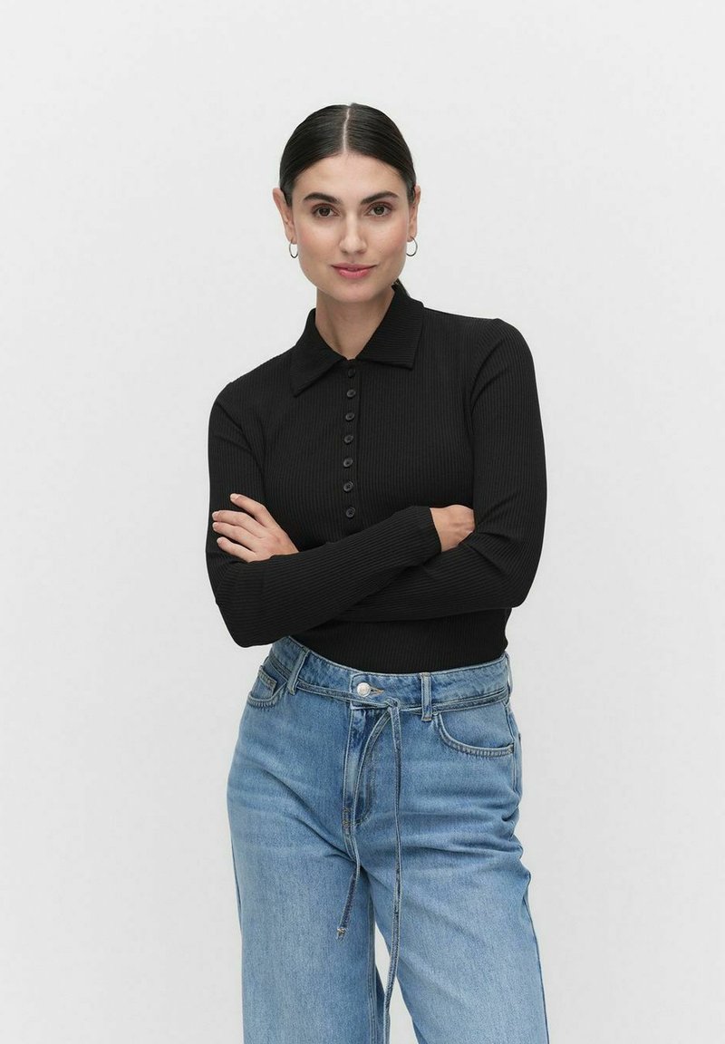 Musta ribbed pitkähiha polo paita nappisuljenta edessä, yhdistettynä vaaleansinihin korkeavyötäröisiin farkkuihin. Malli seisoo kädet ristissä.