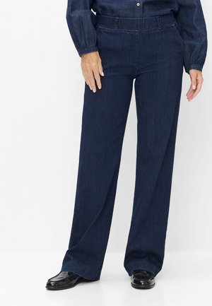 Personne portant un pantalon large en jean bleu foncé avec une taille élastique et des chaussures noires, debout devant un fond blanc.