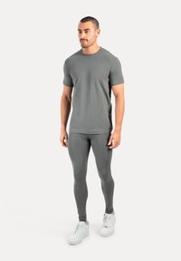 Homme portant une chemise gris ajustée à manches courtes, un legging assorti et des baskets blanches de sport, debout devant un fond clair uni.