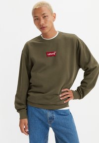 Olivengrønn hettegenser med en rød brodert Levi's-logo på brystet, kombinert med lyseblå jeans. Mykt, strukturert stoff og avslappet passform.