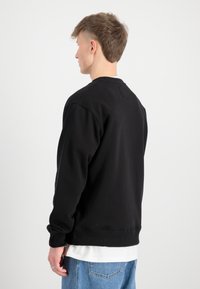 Crni dukserica s rebrastim manžetama i donjim rubom, okruglim izrezom i teksturiranom tkaninom. Nosena je preko bijele majice i uparen je s plavim trapericama.