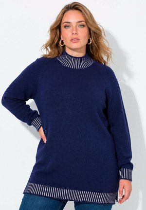 Vrouw met golvend blond haar, die gouden creolen draagt en een marineblauwe trui met witte gestreepte manchetten, zoom en kraag, poserend tegen een witte achtergrond.