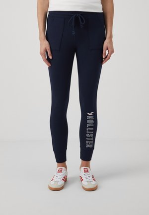 Osoba w granatowych spodniach dresowych z "Hollister" na lewej nogawce, białej koszuli i białych butach Adidas z czerwonymi paskami, stojąca w pomieszczeniu.