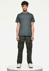 Joven de pie en una plataforma redonda, vistiendo pantalones cargo verde oscuro, camiseta de manga corta gris oscuro y zapatillas blancas, contra un fondo liso.