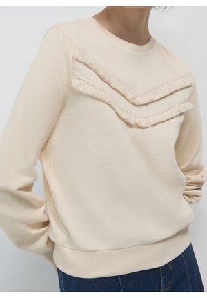 Personne portant un sweat-shirt crème avec des détails chevrons à franges sur la poitrine et un jean bleu, bras détendus sur les côtés.