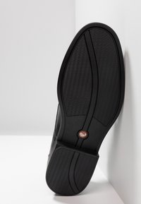 Semelle d'une chaussure habillée noire avec une surface texturée antidérapante, des lignes de design incurvées et un petit logo rond près de la région de la voûte plantaire.