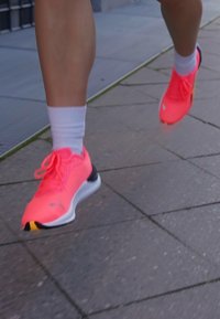 Zapatos deportivos de color rosa brillante con detalles en negro y amarillo, con un diseño ligero y cordones visibles, usados con calcetines blancos sobre pavimento.