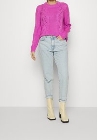 Person som bär en ljusrosa stickad tröja, ljusblå högmidjade jeans och krämfärgade klackade ankelstövlar, står mot en neutral bakgrund.