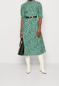 Zielono-biała sukienka w kwiaty z paskiem w talii, krótkimi rękawami i długością do połowy łydki, połączona z białymi botkami do kolan i czarną torebką.