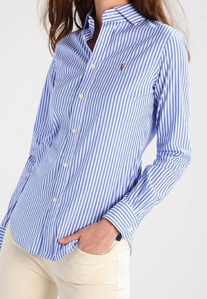 Vrouw met een blauw-wit verticaal gestreept overhemd met kraag en knopen, hand in de zak, gecombineerd met lichtbeige broek.