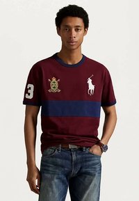 Νεαρός άνδρας που φοράει ένα μπορντό πόλο μπλουζάκι με ναυτικό ρίγες, λευκό λογότυπο παίκτη πόλο, αριθμό 3 στο μανίκι, κεντημένο έμβλημα και μπλε τζιν.