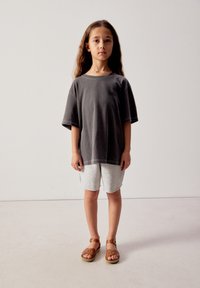 Jeune fille aux longs cheveux, vue de face, portant un t-shirt gris foncé surdimensionné, un short gris clair et des sandales marron.