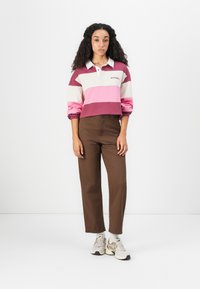 Kvinne med langt krøllete hår iført en stripete rugbyshirt, høy midje brune bukser, hvite sokker og beige joggesko som står foran en hvit bakgrunn.