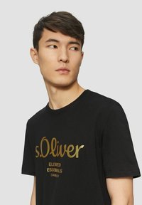 Black cotton t-shirt featuring a large gold printed logo reading "s.Oliver" and "Elevated Essentials" below. Standard crew neck design.
