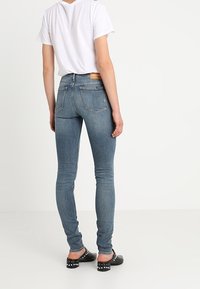 Vaqueros pitillo en denim azul medio con un acabado desgastado, que cuentan con una cintura de talle medio, bolsillos traseros y detalles de costura. Combinados con una camiseta blanca.