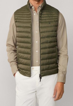 Homme portant une veste matelassée vert olive par-dessus une chemise beige boutonnée et un pantalon blanc, mains partiellement dans les poches, fond neutre.