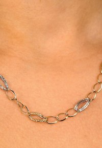Halskette aus Gold- und Silberfarben mit abwechselnd strukturierten und glatten ovalen Formen, die auf einem neutralen Hautton liegt.