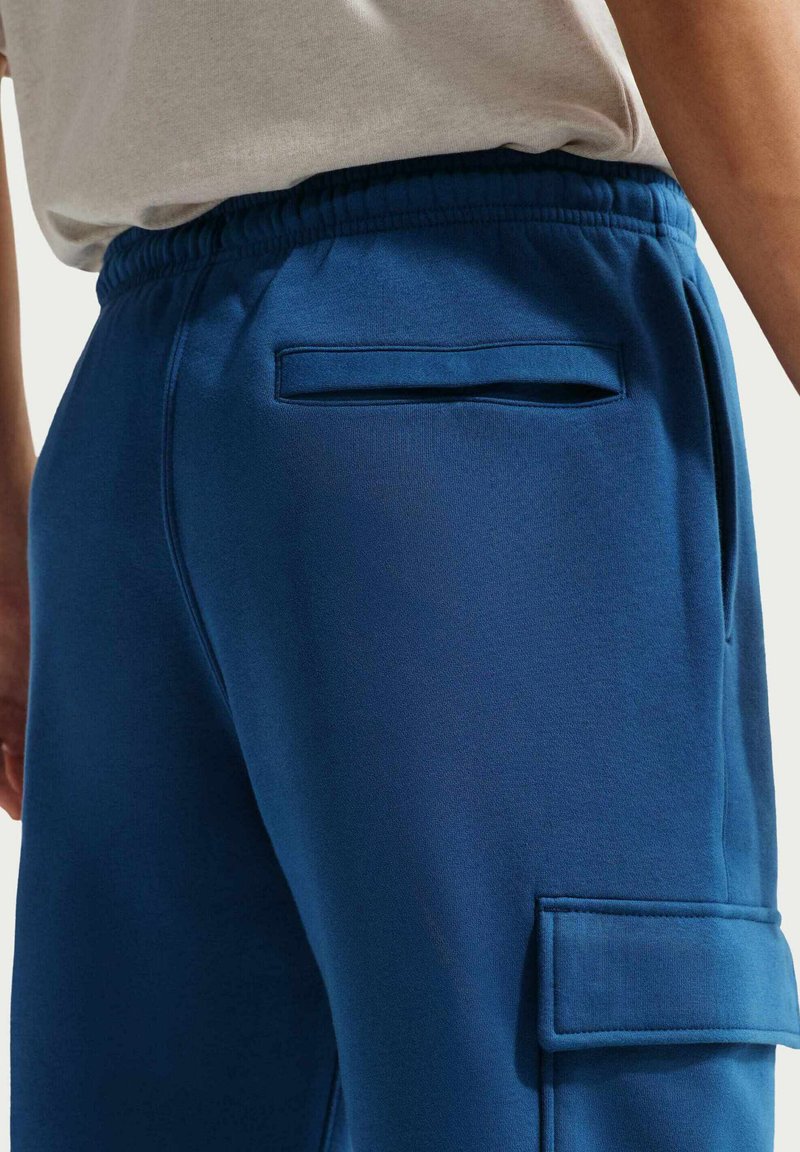 Le pantalon de jogging bleu présente une texture lisse, une ceinture élastique et une poche arrière avec une ouverture rectangulaire, conçus pour le confort et la fonctionnalité.