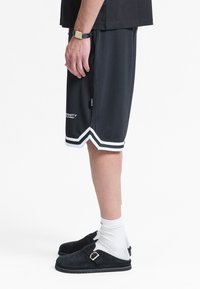 Personne portant un short de sport noir à rayures blanches, des chaussures noires sans lacets avec boucles, des chaussettes blanches et un t-shirt noir à manches courtes.