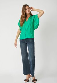 Zelená, voľná blúzka s výstrihom do V a detailom so šnúrkou, spárovaná s širokými temnými džínsami a čiernymi opätkami ozdobenými mašľami.