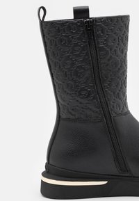 Bottine noire à la cheville avec une finition texturée et embossée. Dispose d'une fermeture éclair latérale, cuir lisse, et un accent métallique discret sur la semelle.