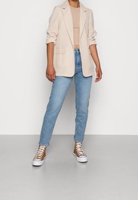 Person som står iklädd en beige rutig kavaj över en beige kort topp, ljusblå jeans och bruna hög-topps sneakers med vita snören.