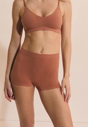 Frau trägt braunen nahtlosen Bralette und hoch taillierte Boyshort-Unterwäsche, steht vor neutralem Hintergrund.