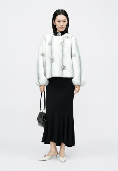 Valge kunstkarusnahast jakk hõbedaste kaunistustega, kombineeritud musta pika seeliku ja kreemjade teravnurksete mules-kingadega, käes must tekstuuriga käekott.