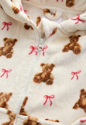 Jachetă din fleece cu fermoar, având un fundal crem cu un model de ursuleți de pluș și fundițe roșii. Textură moale cu un design confortabil.