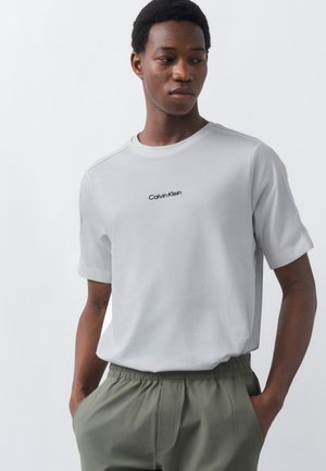 Fiatal férfi világosszürke Calvin Klein pólóban és olívazöld nadrágban, zsebre tett kézzel áll, balra néz.