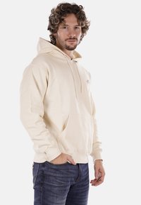 Felpa crema realizzata in morbido tessuto, con tasca a marsupio, cappuccio con coulisse e logo discreto sul petto, abbinata a jeans in denim scuri.