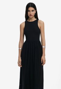 Vestido maxi negro con un cuerpo acanalado, diseño sin mangas y falda fruncida. Presenta una pulsera de puño plateada en el brazo izquierdo.