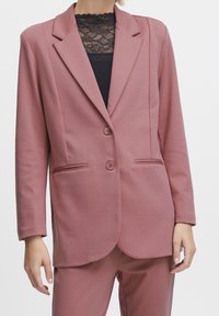 Rosa kavaj med klassisk slag, två knappar och sidofickor i ett mjukt tyg. Bärs över en svart spetsblus för kontrast.