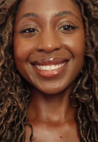 Frau mit dunklem, lockigem Haar, die minimal Make-up trägt. Sie hat ein warmes Lächeln, glänzende Lippen und eine glatte, braune Haut. Klare, helle Augen.
