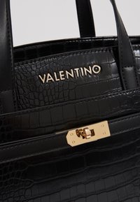 Černá kabelka z krokodýlí kůže s embosovaným vzorem, se zlatým uzávěrem na zámek a logem "Valentino" napsaným zlatým písmem na přední straně.