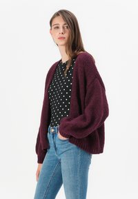 Burgundská pletená cardigan s širokými rukávy přes černou halenku s puntíky, spárovaná s modrými úzkými džíny. Jednoduchý, texturovaný design.
