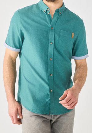 Man wearing a teal short-sleeve button-up shirt with a chest pocket and light gray pants, standing against a plain background.