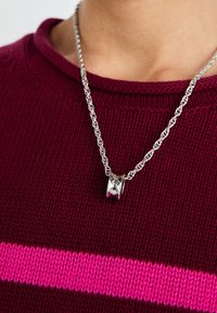 Collier en chaîne torsadée argent avec un pendentif poli en forme de baril argenté porté sur un pull bordeaux avec une bande rose horizontale.