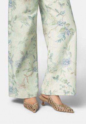 Pies con sandalias doradas de tacón bajo con tiras abiertas, combinadas con pantalones de pierna ancha con un suave patrón floral en tonos pasteles.