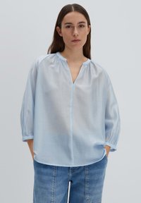 Blusa azzurro chiaro, semi-trasparente, con scollo a V, maniche arricciate e vestibilità ampia. Abbinata a pantaloni corti in denim, caratterizzati da una texture liscia.
